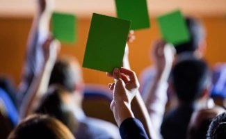 Una reunión de vecinos enfocada desde el fondo de la sala, se observan varias filas de vecinos participando en una votación.