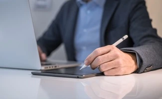 Primer plano de una mano usando un lápiz digital sobre una tableta, con un ordenador portátil abierto en el escritorio.