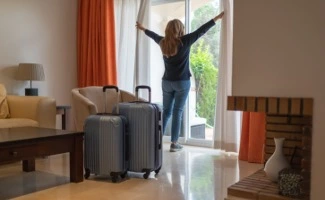 Persona en un alojamiento turístico con maletas, abriendo las cortinas y mirando al exterior, representando una estancia temporal o alquiler vacacional.