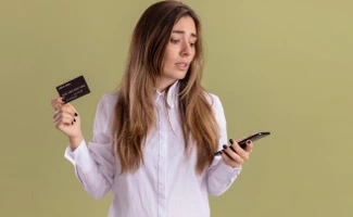 Mujer sosteniendo una tarjeta bancaria y mirando su teléfono móvil con expresión de preocupación, simbolizando gastos, pagos o dificultades económicas.