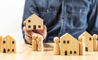 Mano sosteniendo una pequeña casa de madera sobre una mesa con figuras de personas y otras casas de madera, representando una decisión o elección relacionada con vivienda o propiedad.